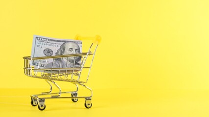 Shopping cart and one hundred dollar bill on a yellow background, closeup. Black Friday Shopping and Discount Concept