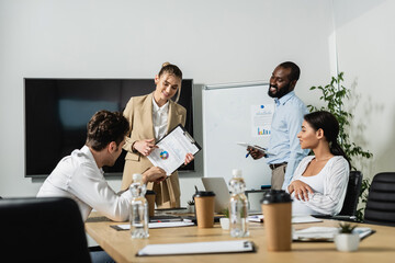 smiling businesswoman pointing at graphs while talking to multiethnic coworkers