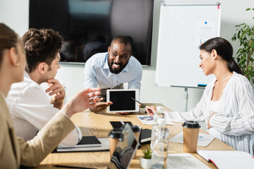 smiling african american businessman showing digital tablet with blank screen during meeting with colleagues