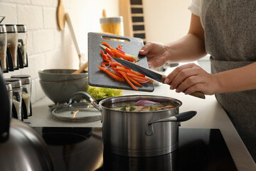Woman putting cut vegetables into pot with delicious bouillon on stove in kitchen, closeup - Powered by Adobe