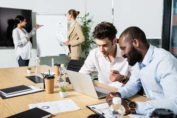 interracial businessmen looking at laptop near blurred businesswomen near flip chart