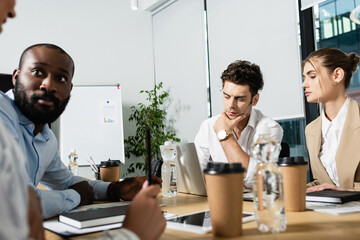 young businessman thinking near laptop and blurred interracial friends