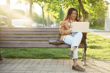 African american woman freelancer sitting on a bench in a street and using laptop - Powered by Adobe