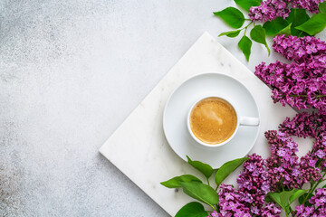 White cup of fresh coffee on marble tray and purple lilac with green leaves on grey textured background. Morning cappuccino and spring flowers. Blogging concept. Flat lay, top view, copy space.