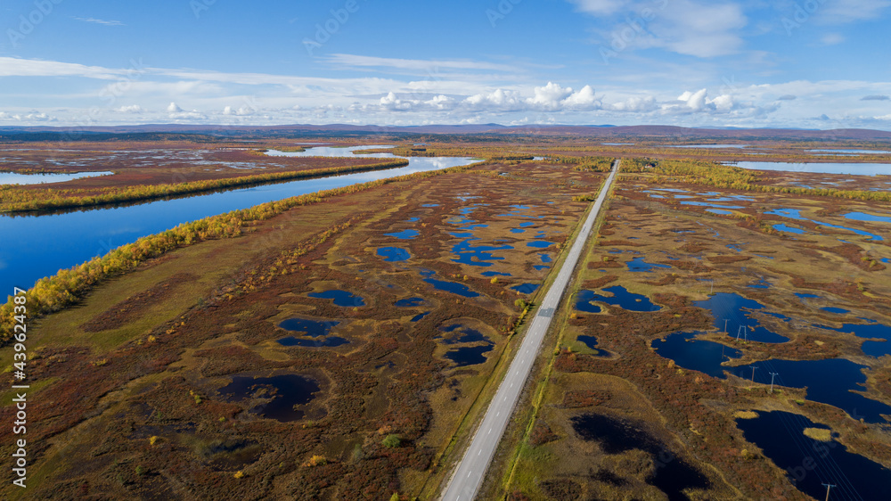 Wall mural Aerial view of asphalt forest road, river and lakes in Lapland, highway and colourful autumn foliage. Brown, yellow autumn colors. Blue sky and clouds. - Wall murals