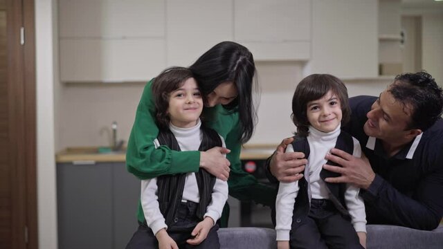 Smiling parents walking to absorbed twin brothers watching TV sitting on couch and hugging adorable sons talking. Happy multiethnic family resting on weekend at home indoors