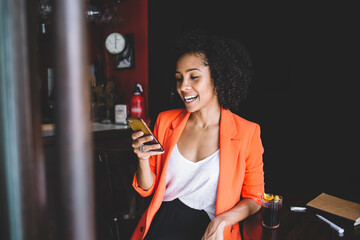Naklejka na ściany i meble Cheerful black woman using smartphone in cafe