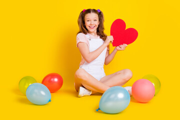 Fototapeta na wymiar Full length body size photo schoolgirl at party with balloons smiling showing red heart isolated bright yellow color background