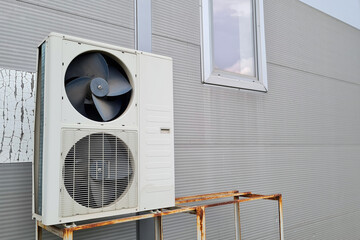 White double outdoor unit of air conditioner on gray wall of shopping center. One of fans without a protective grill, possibly broken.