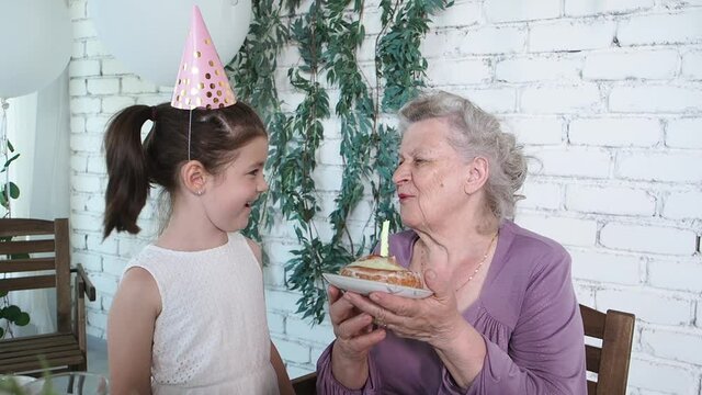 Slow motion. Happy smiling little cute girl and senior grandmother celebrate 85th birthday, holds cake and blowing out candle together, white brick background. Enjoy, respect and warm relation concept