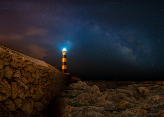 Vía láctea en Menorca, faro iluminando el cielo, muro de piedra y rocas de acantilado