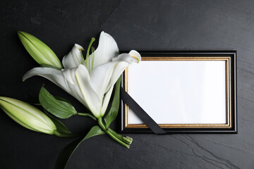 Funeral photo frame with ribbon and white lily on black table, flat lay. Space for design