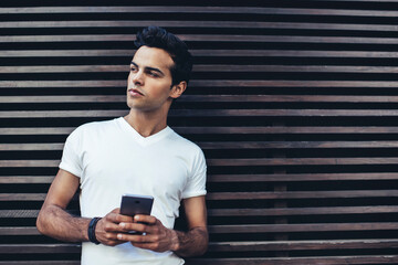 Pondering male blogger with smartphone technology thoughtful looking away at publicity area, Caucasian hipster guy dressed in white t shirt with copy space area for label name using mobile device