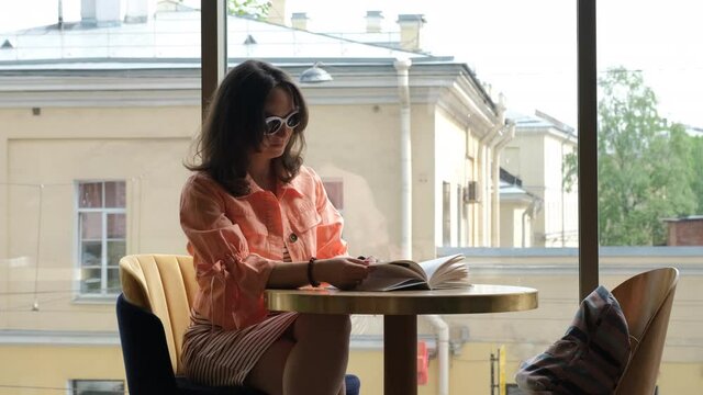 Young Female in Sunglasses Reading a Book while Sitting at Table in Cafe. Leisure, Literature, Hobby and People Concept