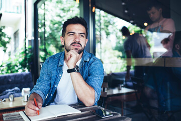 Serious male student with education textbook learning in street cafeteria thinking about...