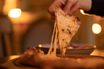 A woman's hand takes a slice of hot vegetarian four cheese pizza with stretched cheese close-up, warm evening light. Freshly prepared Italian dish in the restaurant, cafe