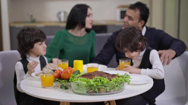 Dinner table with blurred family eating food at background talking smiling. Positive Caucasian woman Middle Eastern man and twin brothers at home in the evening. Family