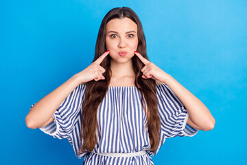 Fototapeta premium Photo of childish funny young woman dressed striped outfit press fingers bloated cheeks isolated blue color background