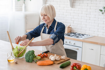 Cheerful old senior elderly aged woman adding tomatoes to the vegetable salad, fresh ingredients, veganism and vegetarian concept. Cooking at home. Grandmother preparing food in the kitchen