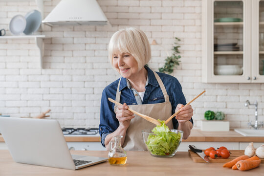 Old senior aged grandmother woman looking for recipe on laptop cooking preparing salad, healthy food and meal. Fresh ingredients, cooking at home. Vegetarian and veganism