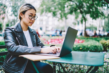 Japanese female sitting at street cafe with laptop computer doing remote job, youthful IT developer in optical eyewear typing and searching information from social networks during freelance work