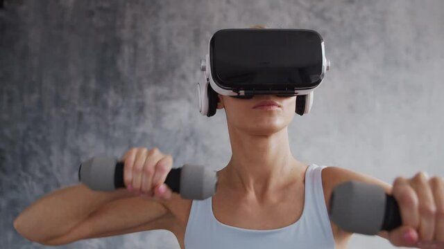 Young and sporty girl in sportswear is doing exercises at home using virtual reality helmet. Fit and slender blond woman goes in for sports and fitness. Healthcare, fat burn and wellness.