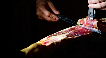 Gastronomía y alimentación en España. Detalle de primer plano de maestro cortador de jamón ibérico sobre fondo oscuro.