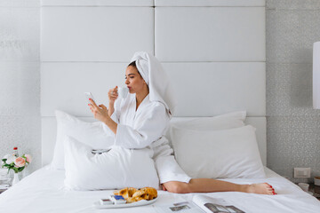 Young woman wearing bathrobe, drinking coffee and using mobile phone at bed in hotel room - 439638129