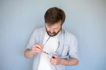 Concentrated Caucasian man cleans glasses from dust and dirt, wiping them with his shirt. Gray...