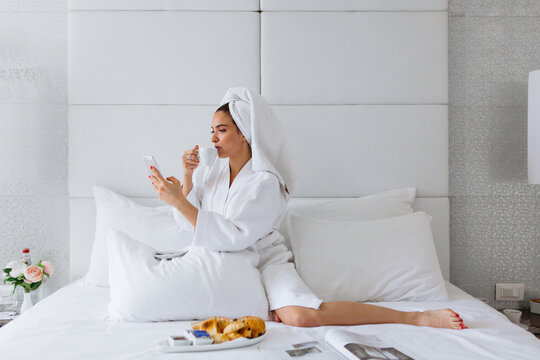 Young woman wearing bathrobe, drinking coffee and using mobile phone at bed in hotel room