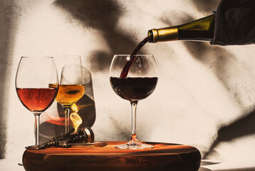 Wine tasting. Red wine pouring into glass on background with selection of red, white and rose wines in glasses. Hard sunlight and shadows from foliage - obrazy, fototapety, plakaty
