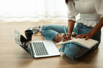 Naklejka premium Close-up of a woman sitting on the floor holding a pencil and taking notes using a laptop at home.