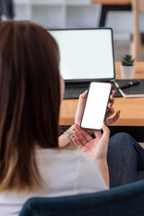 Back view of woman holding a smartphone blank white screen sitting at the office. Mock up.