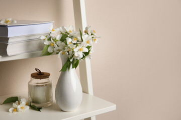 Bouquet of beautiful jasmine flowers in vase and candle on shelving unit near beige wall indoors,...