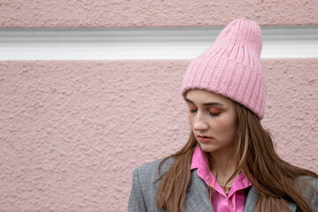 Portrait of a teenage girl with closed eyes dressed in a coat, a pink shirt and a knitted hat standing on a blurred background of a textured wall. A girl with fatigue on her face sleeps on the go.