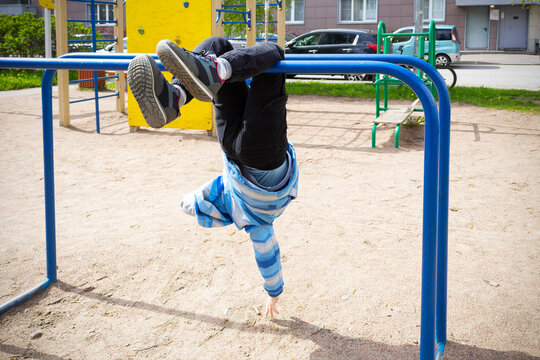 Child in blue jacket hanging on crossbars upside down in playground, against backdrop of house and cars.