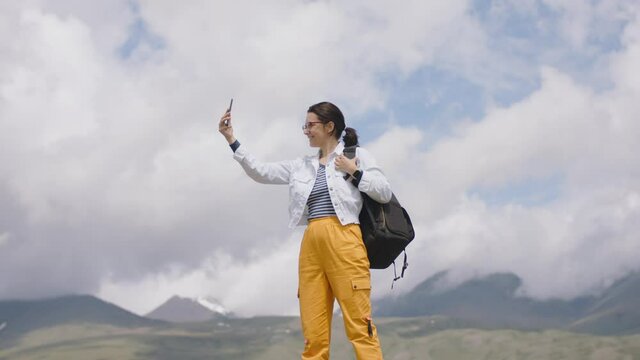 Travel blogger takes selfie on smartphone camera in mountains. Tourist takes pictures with mobile phone camera. Young woman traveler with backpack shoots video