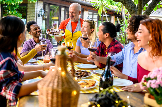 Group of happy friends drinking red wine in a garden while waiter is serving them grilled meat - Happy friends eating bbq food at restaurant - Life style concept about friendship and happiness