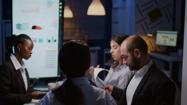 Selective focus workaholics focused multi-ethnic businesspeople overworking in company office meeting room brainstorming ideas late at night. Coworkers sitting at desk analyzing statistics paperwork
