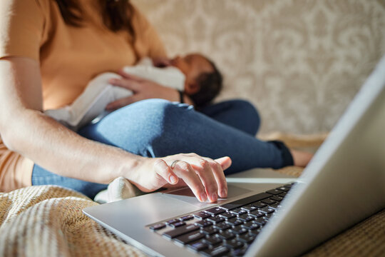 Mother multi-tasking, holding baby infant and using computer laptop at home. 