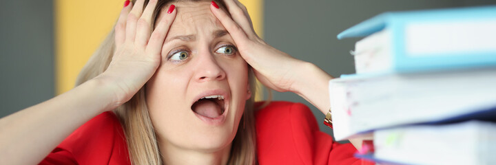 Woman holding her head and is surprised in front of many folders on table - obrazy, fototapety, plakaty