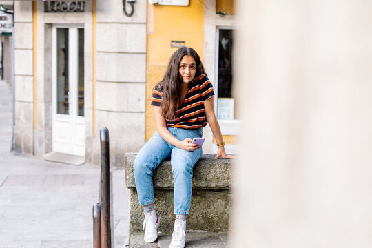 Young woman sitting on a wall while holding a phone at downtown of cute town
