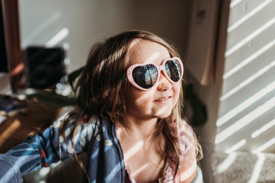 Preschool age girl with pink heart sunglasses and princess dress standing in window light.