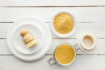 Washing the dishes with mustard powder concept. White clean plates with bowl and jar of yellow mustard powder and wooden dish washing brush and class jar container with natural washing brush.