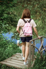 a young brunette with pink backpack on her back stands on a bridge by the river. a white-and-red dog runs around nearby. Rear view