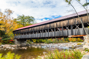 Old wooden covered bridge over a mountain river on a cloudy autumn day. Beautiful autumn colours. White mountain National Forest, NH, USA.