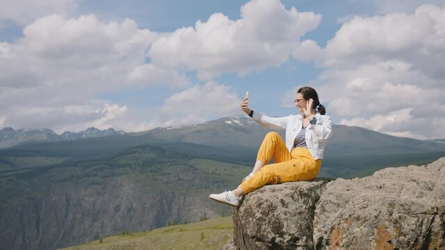 Woman tourist makes selfie on smartphone camera while sitting on edge of cliff in mountains. Traveler video call and shares his impressions with friends