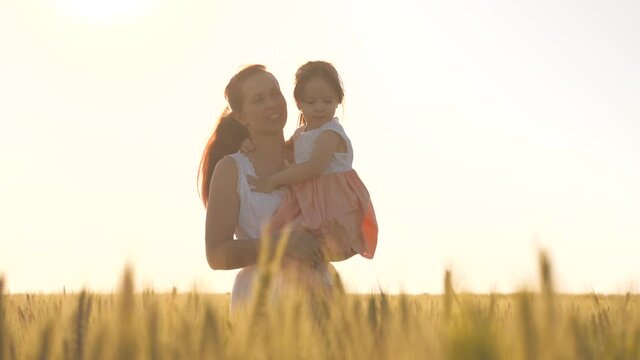 Baby is in mom's arms. Mom and little daughter are walking in a green wheat field, hugging and kissing. A child and mom are walking in wheat field. Happy family travel. Farmer woman and child in field