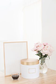 Vertical frame and gift box mockup on a wooden table in the kitchen. Glass jug with a bouquet of pink peonies and a cup of black coffee. Scandinavian style interior.