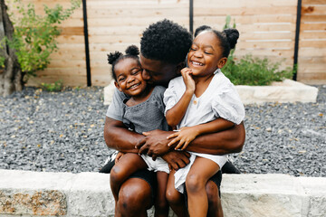 A father sitting outdoors in the backyard having a conversation with his two young daughters & giving them a hug